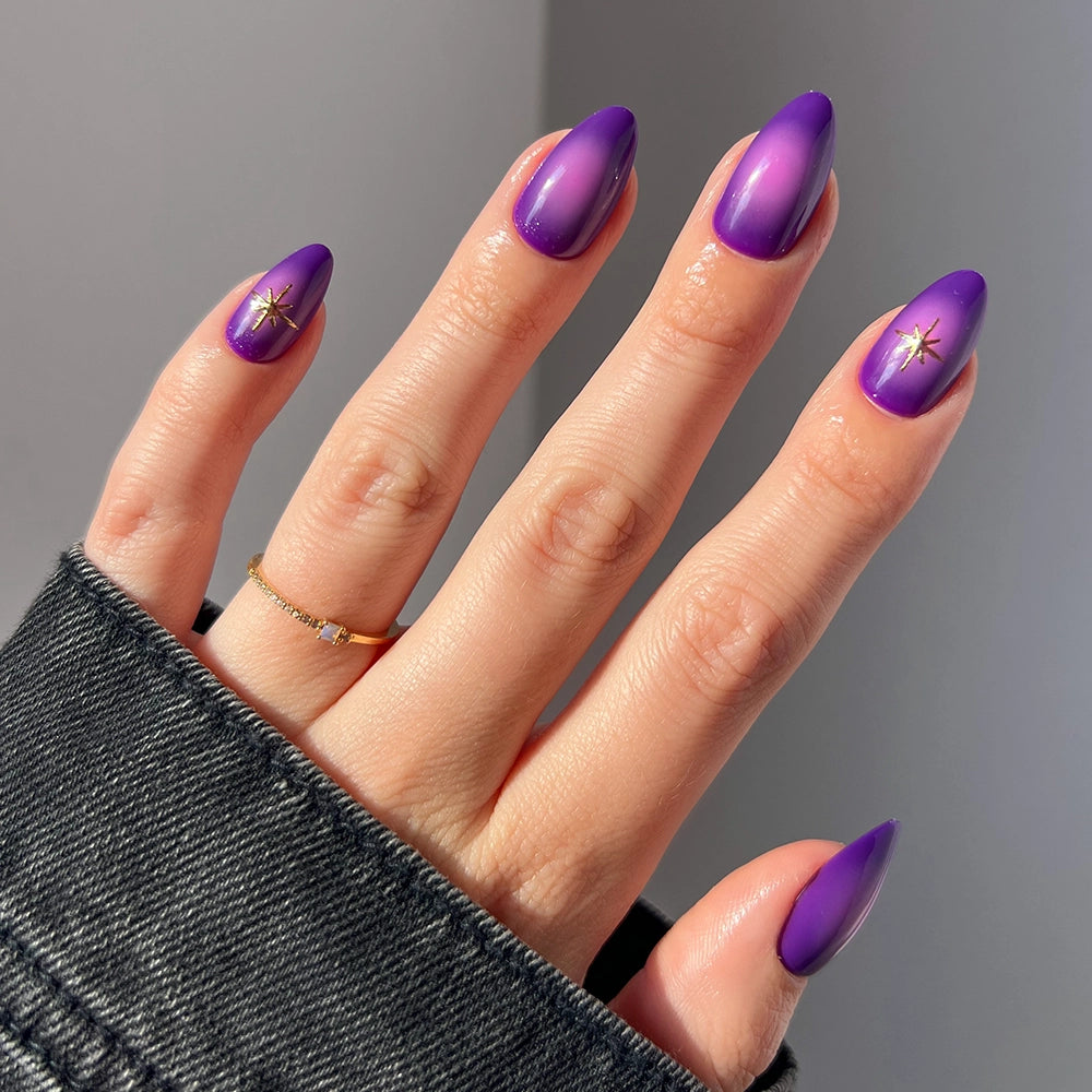 A hand with long, almond-shaped glamnetic Tarot press-on nails painted in gradient purple with a subtle shine. Two nails feature a small gold star design. The person wears a gold ring and a dark denim jacket.