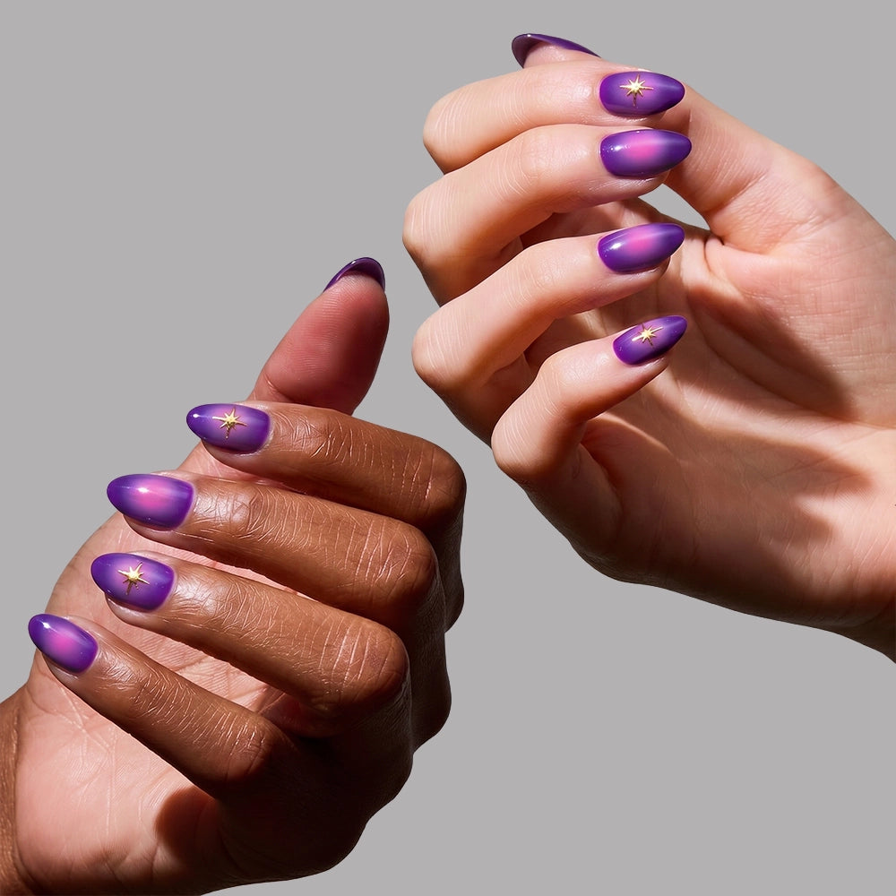 Two hands display glamnetic’s Tarot press-on nails, shaped short almond and painted in a glossy purple gradient with gold star accents. The mystical design stands out beautifully against a plain light gray background. - Product image