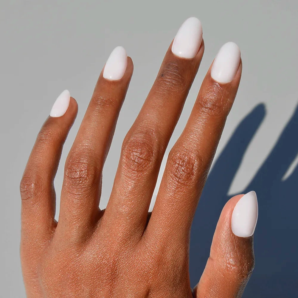 A hand with medium brown skin displays long, almond-shaped nails painted in glossy white "Clean Girl" press-ons by glamnetic, set against a light grey background.
