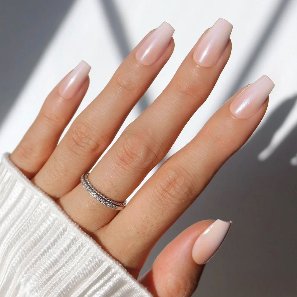 A hand flaunts the "Delicate Touch" coffin nails by glamnetic in pearly pink polish, paired with a double-band silver ring accented with small diamonds and a white, pleated sleeve partially covering the wrist.