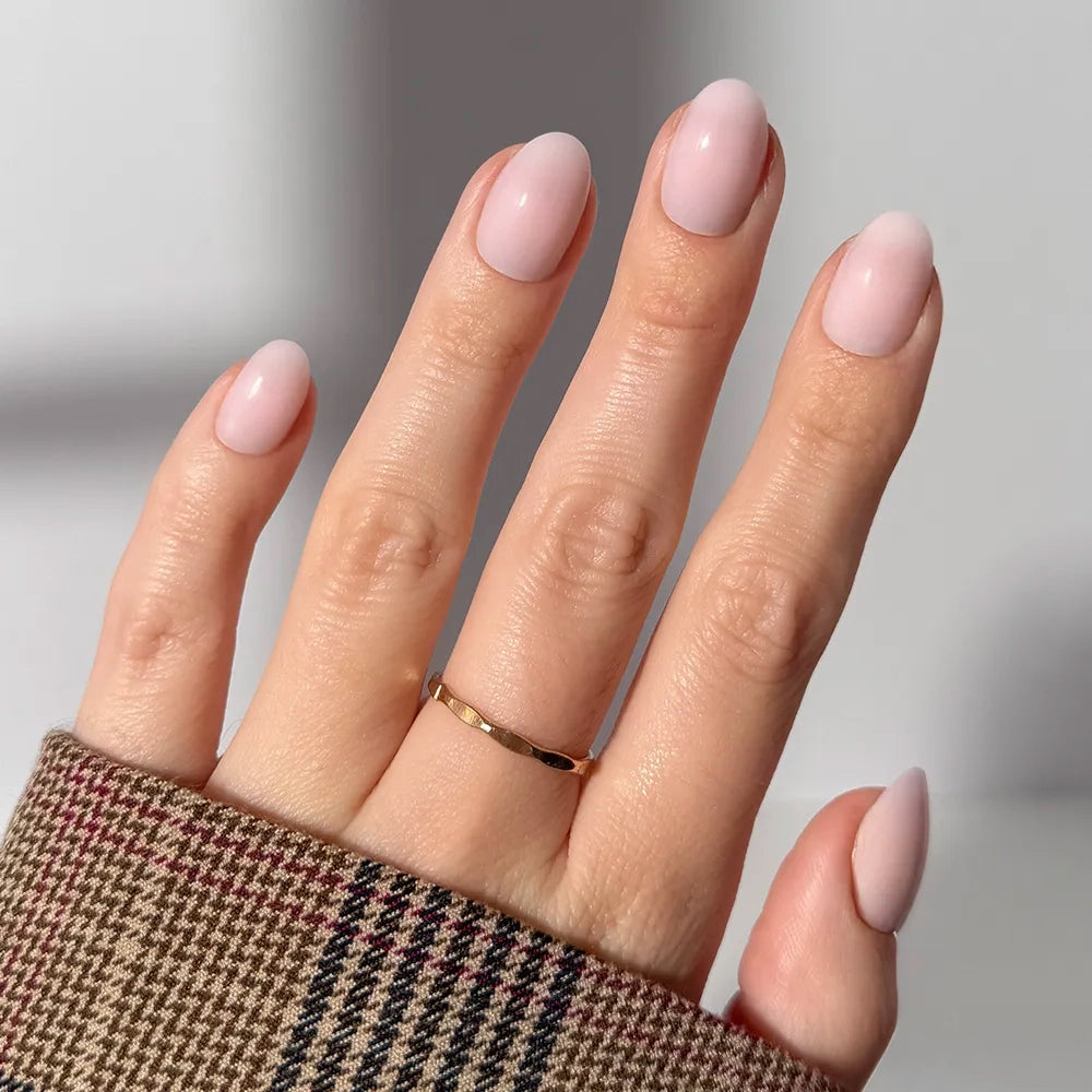 A hand with short, almond-shaped glamnetic “Doll Cheeks” press-on nails in pale pink, wearing a thin gold ring on the middle finger and a brown plaid sleeve partially covering the wrist.