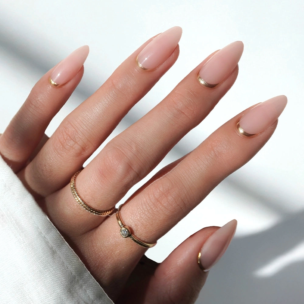 A hand wears glamnetic's Goal Getter almond-shaped nude pink press-on nails with gold cuticle accents, plus two gold rings (a plain band and a diamond ring). The sleeve of a white garment is partially visible.
