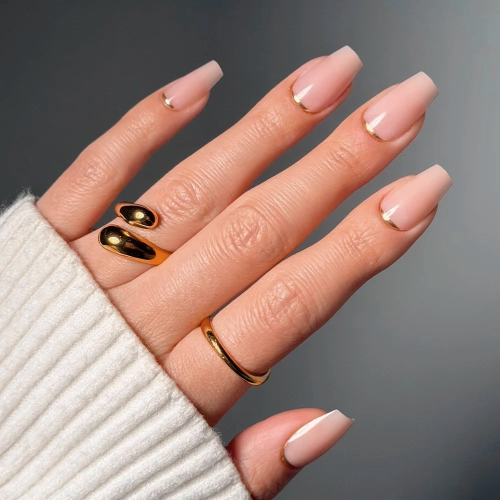 A hand shows off glamnetic’s Gold Goals: long, square-shaped nude pink coffin nails with thin gold tips. Two gold rings and a gold double-finger ring accent the look, partly covered by a cream ribbed sweater sleeve.