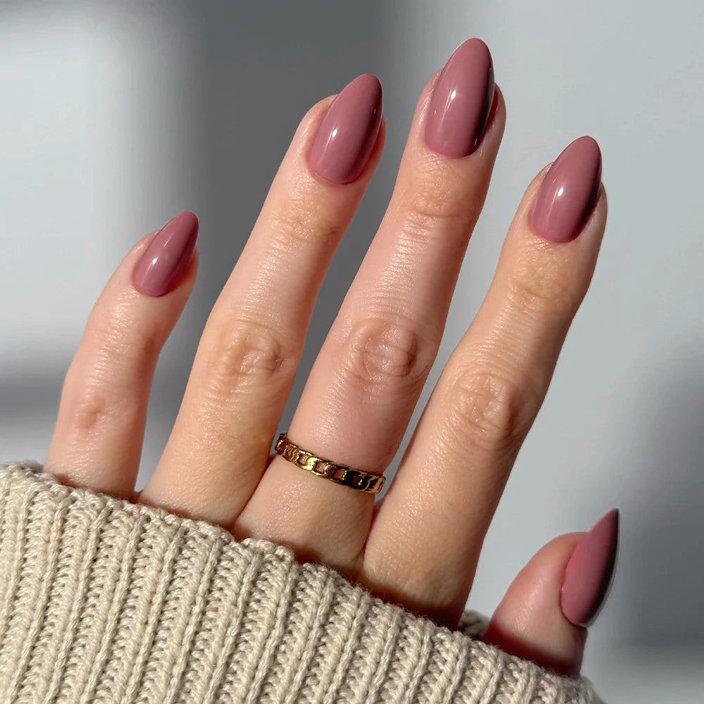 A hand with long, almond-shaped nails in mauve using glamnetic Mauve Obsession press-ons. A gold ring adorns the middle finger, and a beige knit sleeve partially covers the hand.
