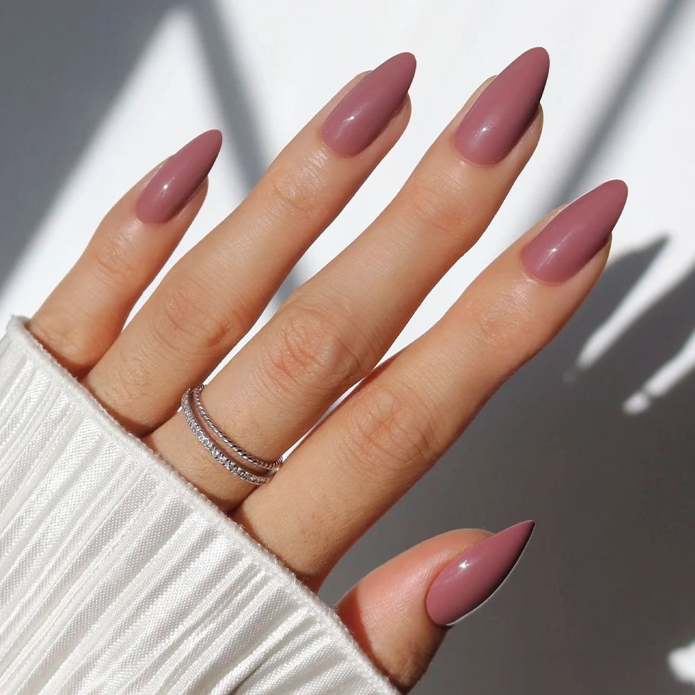 A hand features glamnetic's Mauve Wife long, almond press-on nails in mauve polish, with a double-band silver ring and a white ribbed sleeve partly covering the wrist.