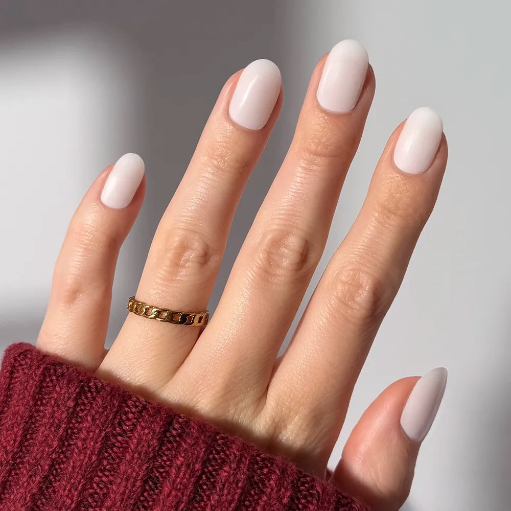 A hand with neatly manicured, almond-shaped glamnetic Milk Bath press-on nails in pale nude, wearing a gold ring on the middle finger and a burgundy knit sweater, shown against a plain light background.