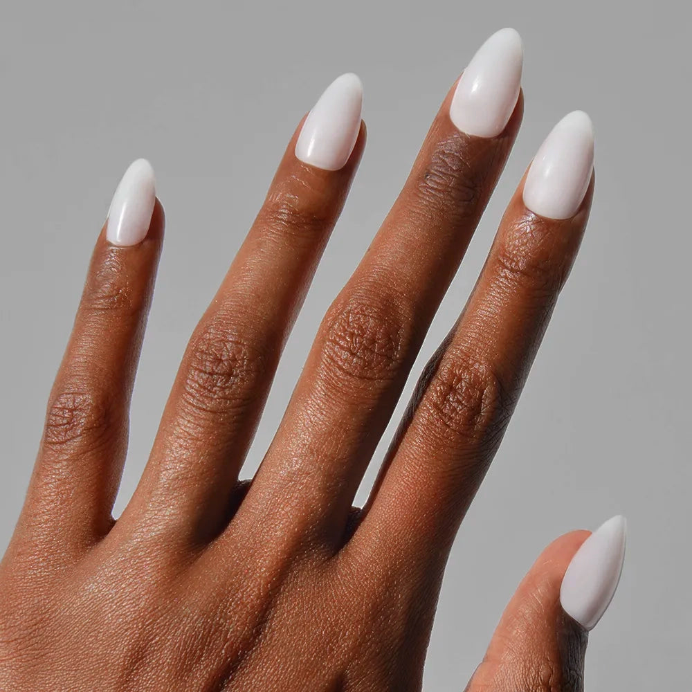A hand with medium brown skin and long, almond-shaped nails wears glossy glamnetic Milky White press-ons, shown against a plain light gray background.