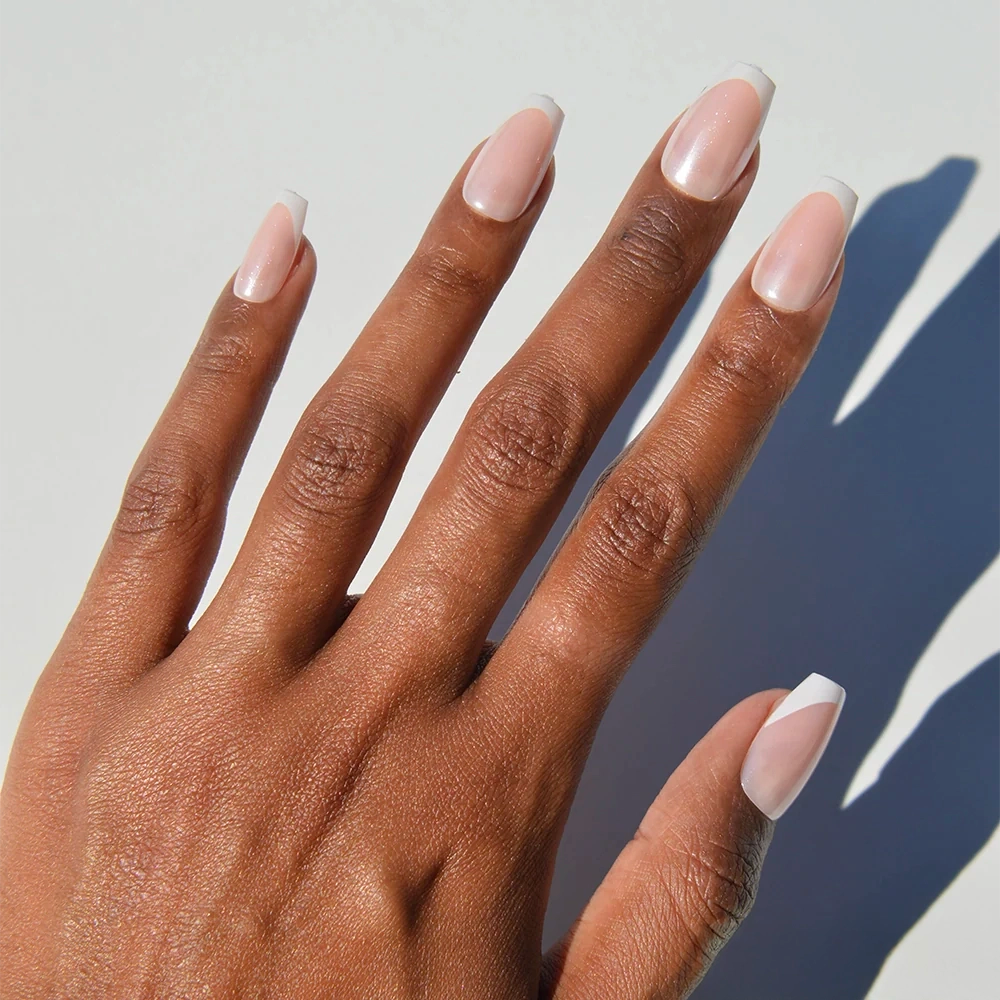 A hand with medium brown skin is shown wearing glamnetic’s Pearl Glaze nails in a modern French tip style—medium coffin-shaped with a light pink base and thin white tips—against a white background.