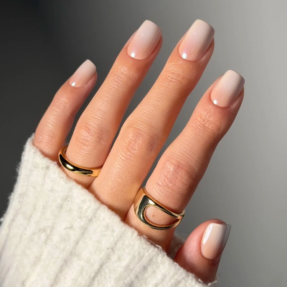 A hand showcases glamnetic’s Sugar Veil nails in a nude ombré, short squoval style. The look is paired with two gold rings and a white ribbed sweater sleeve, set against a neutral backdrop.