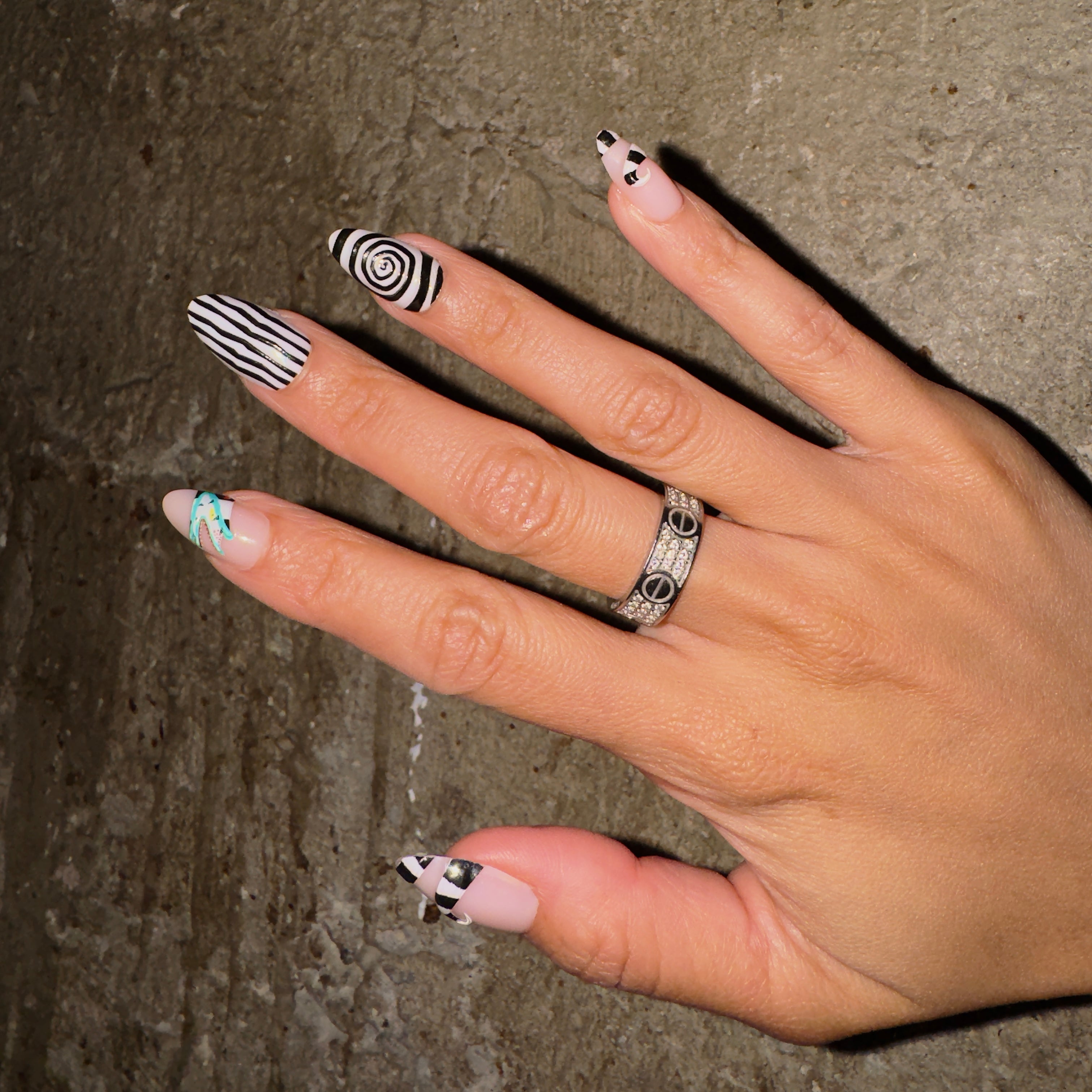 A hand with long nails features bold black and white geometric nail art inspired by the glamnetic Sandworm set. Stripes, swirls, and triangles stand out against textured concrete, while a silver ring adorns the middle finger. - Product image