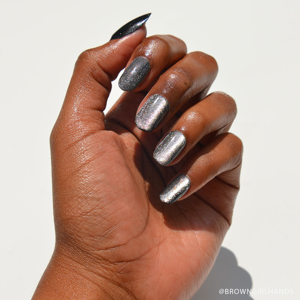 A hand with medium-brown skin displays short, round nails painted in a shimmery pewter shade from glamnetic’s Galactic set, set against a white background. - Product image