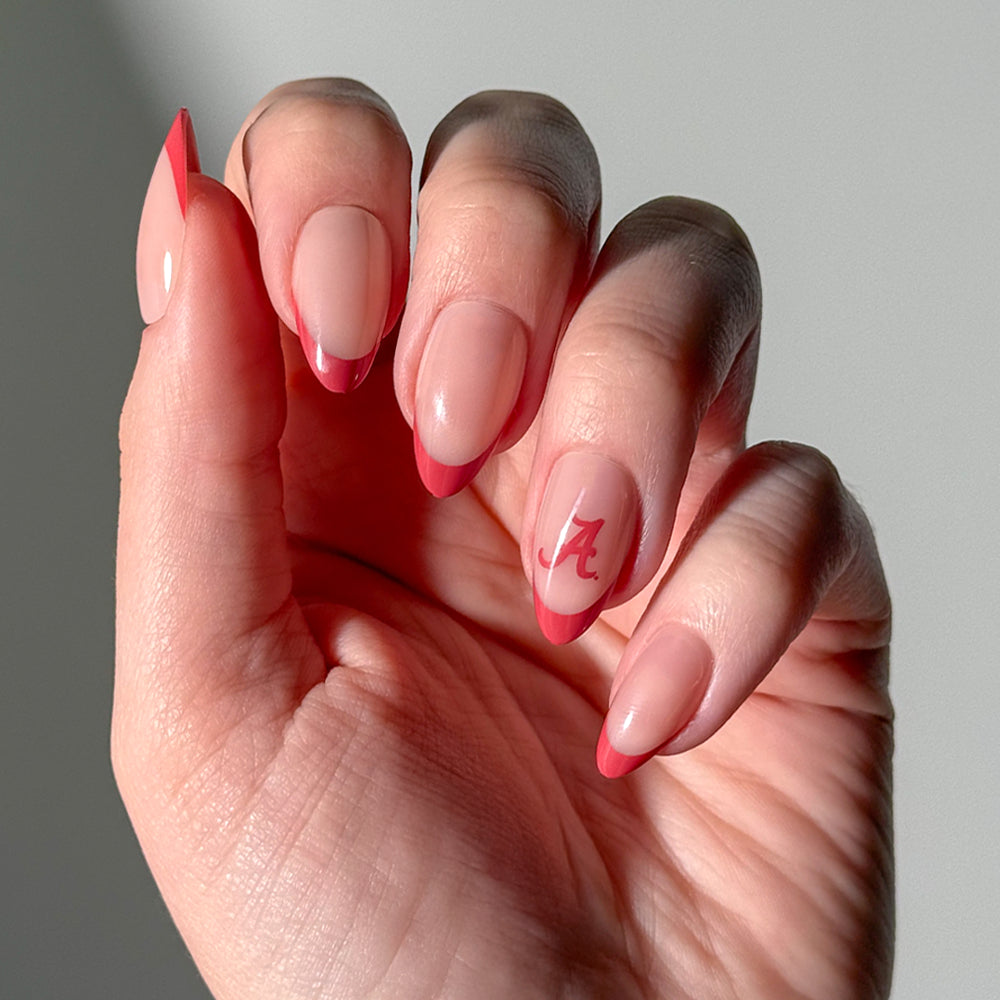 A hand with almond-shaped Collegiate Press-On Nails from glamnetic showcases a pink French tip manicure, with the ring finger featuring a red letter A design.