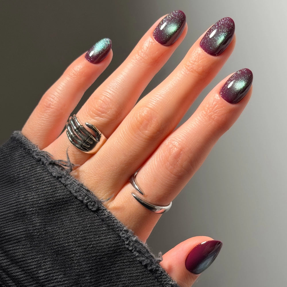 A hand with purple and green shimmery ombre nails, wearing two silver rings, and partially covered by a black denim sleeve. - Product image