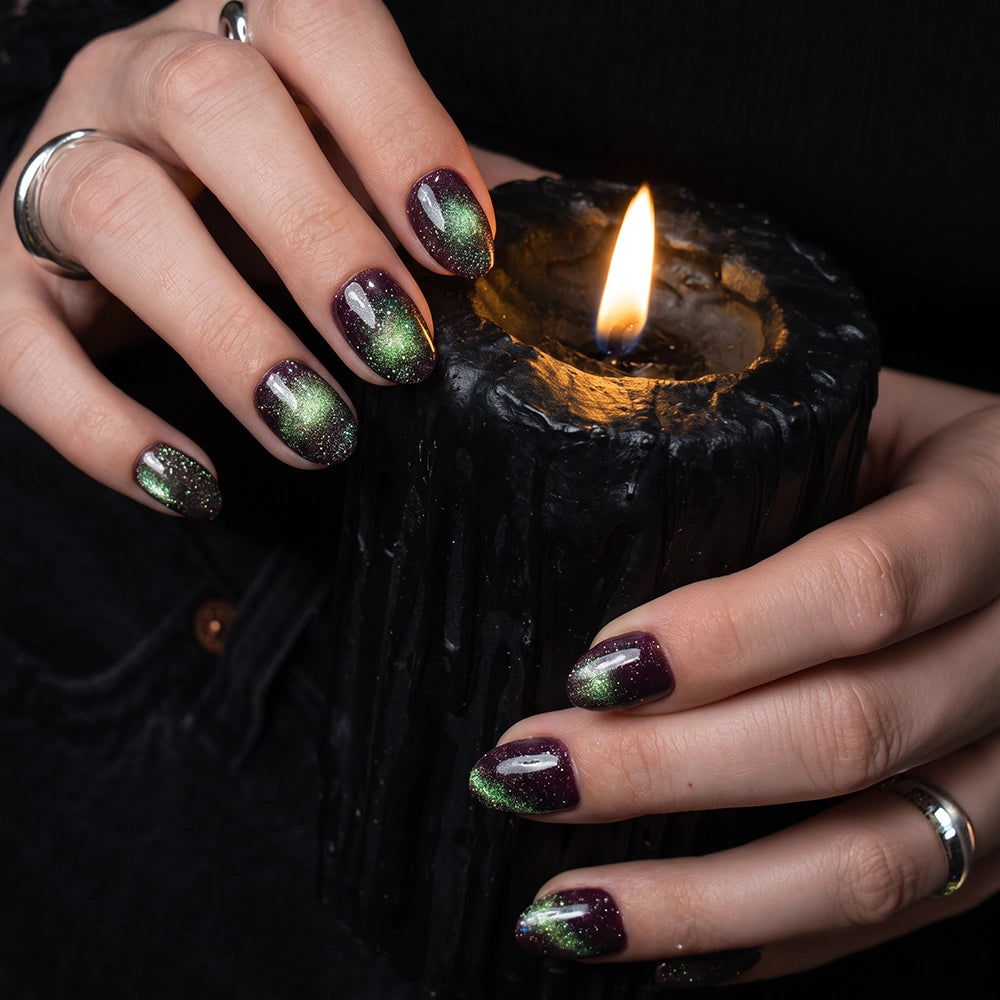 Hands with shiny green and purple ombre velvet finish nails, created with glamnetic’s Alakazam, hold a lit black textured candle. Silver rings and dark clothing add to the moody, gothic atmosphere. - Product image