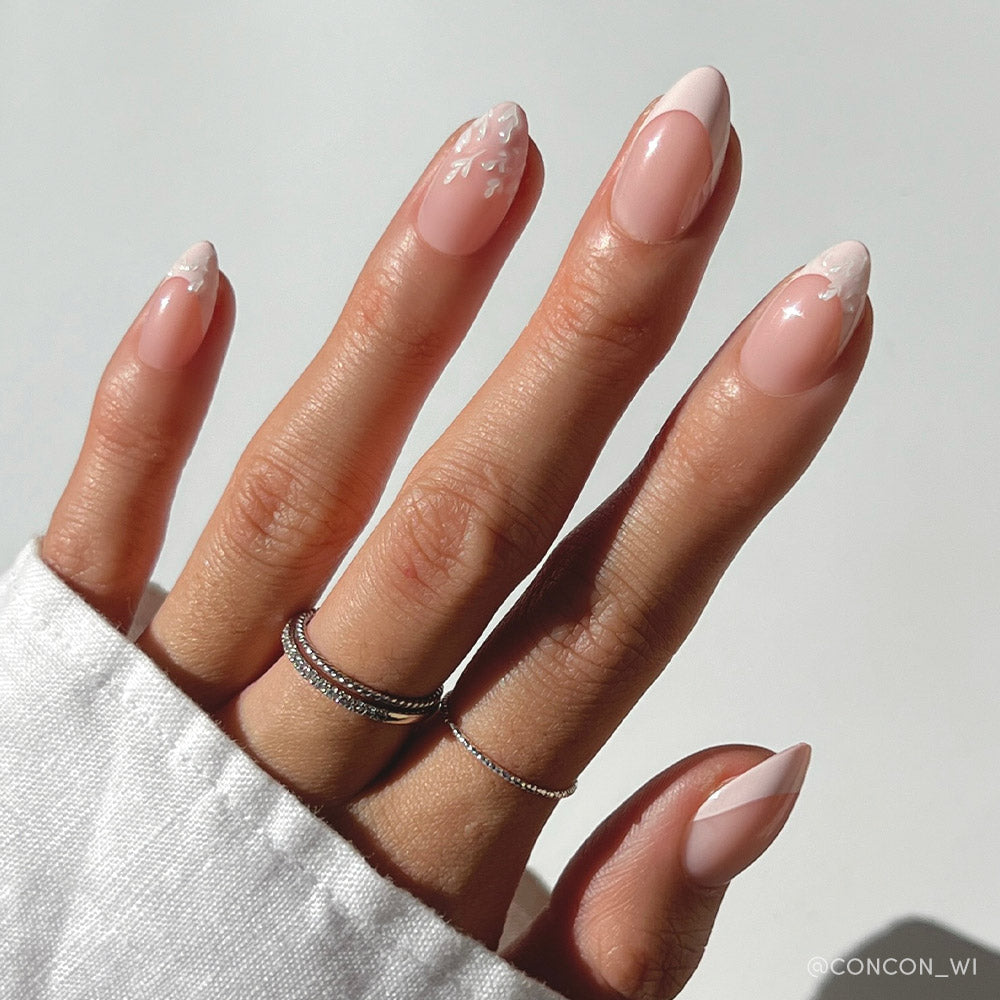 A hand with short almond nails in glamnetics All Time Love: a pink base, delicate French tips, and white floral nail art. The vegan, cruelty-free manicure is styled with two thin silver rings and a white sleeve against a plain background. - Product image