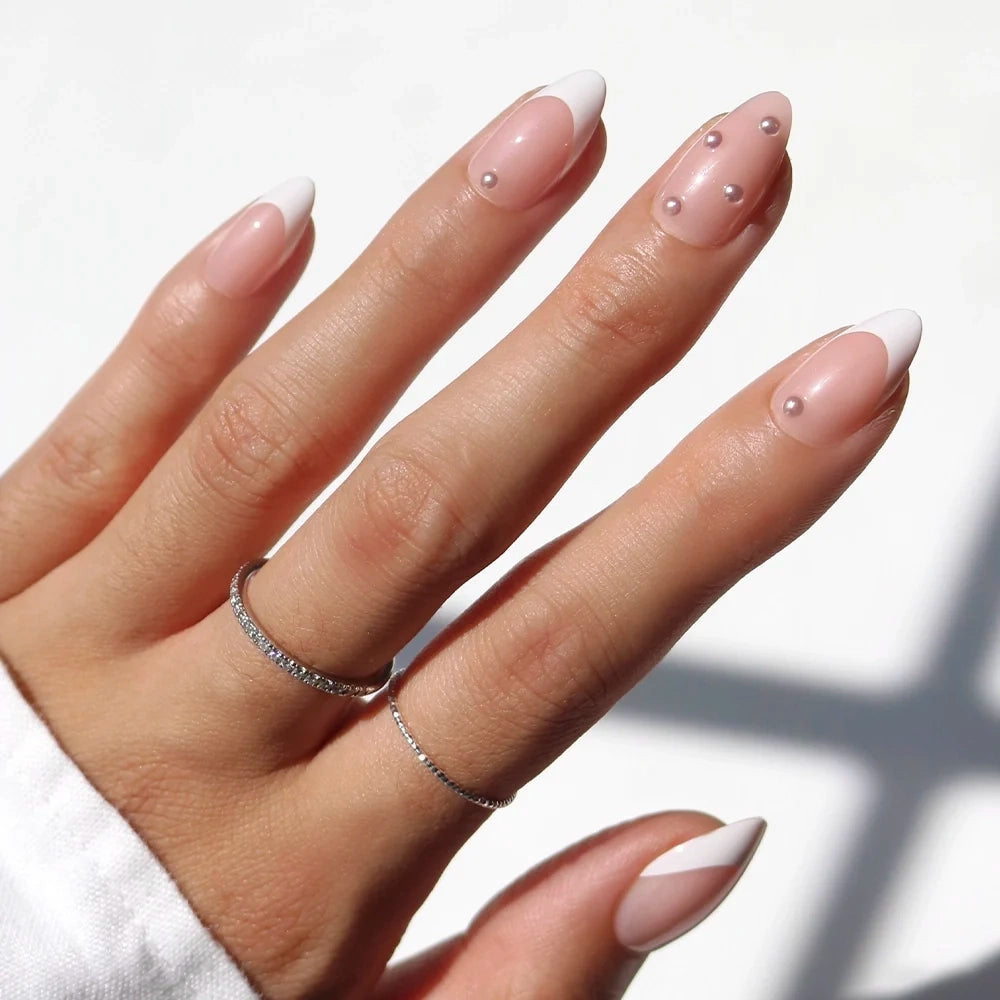 A hand adorned with glamnetic’s ALWAYS & FOREVER nails, featuring almond-shaped French tips and delicate pearl-like accents on two nails. Two thin rings on the middle and ring fingers enhance this timeless and chic pearl nail design.