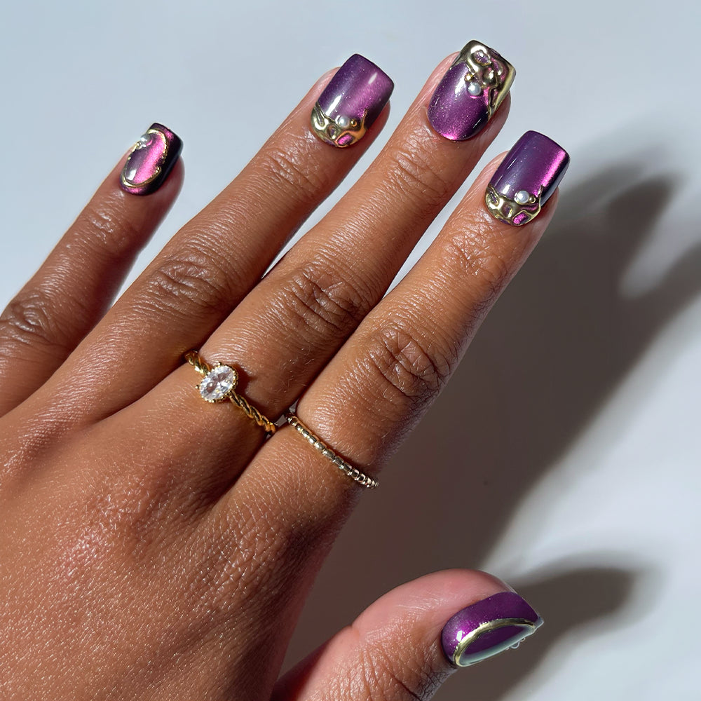 A hand with short squoval nails displays the glamnetic Amethyst Press-On Nails, featuring a subtle velvet purple finish and adorned with elegant gold accents, small pearls, and gold details for a luxurious look. The person accessorizes with two rings: a delicate gold band and another set with a flower-shaped cluster of gems.