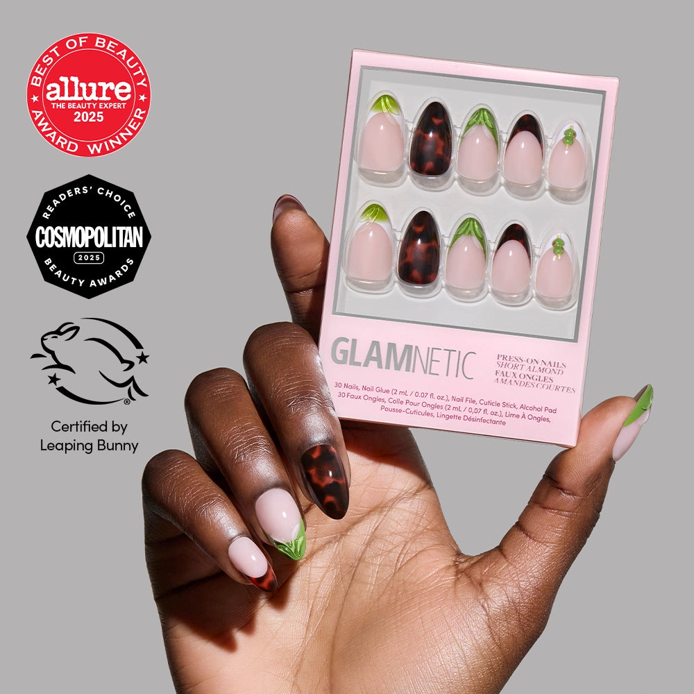 A hand with almond-shaped Antipasto press-on nails by glamnetic, featuring green leaf tips and dark tortoiseshell accents, holds a box of the same nails. Award badges from Allure, Cosmopolitan, and Leaping Bunny appear beside the hand.