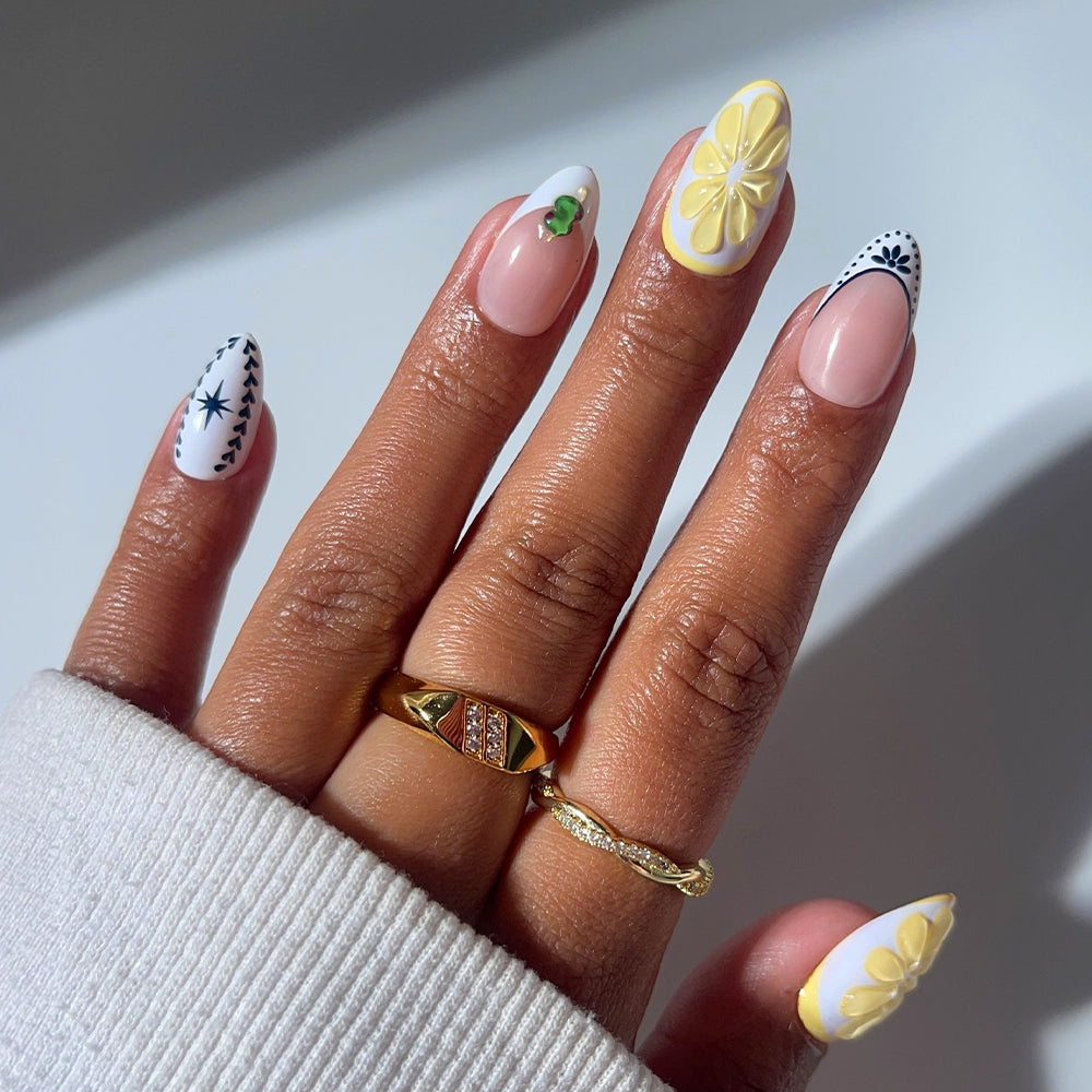 A hand with glamnetic's Aperitivo Hour press-on nails showcases yellow 3D flowers, black and white patterns, a hint of green, gold rings, and a cream sleeve covering part of the hand.