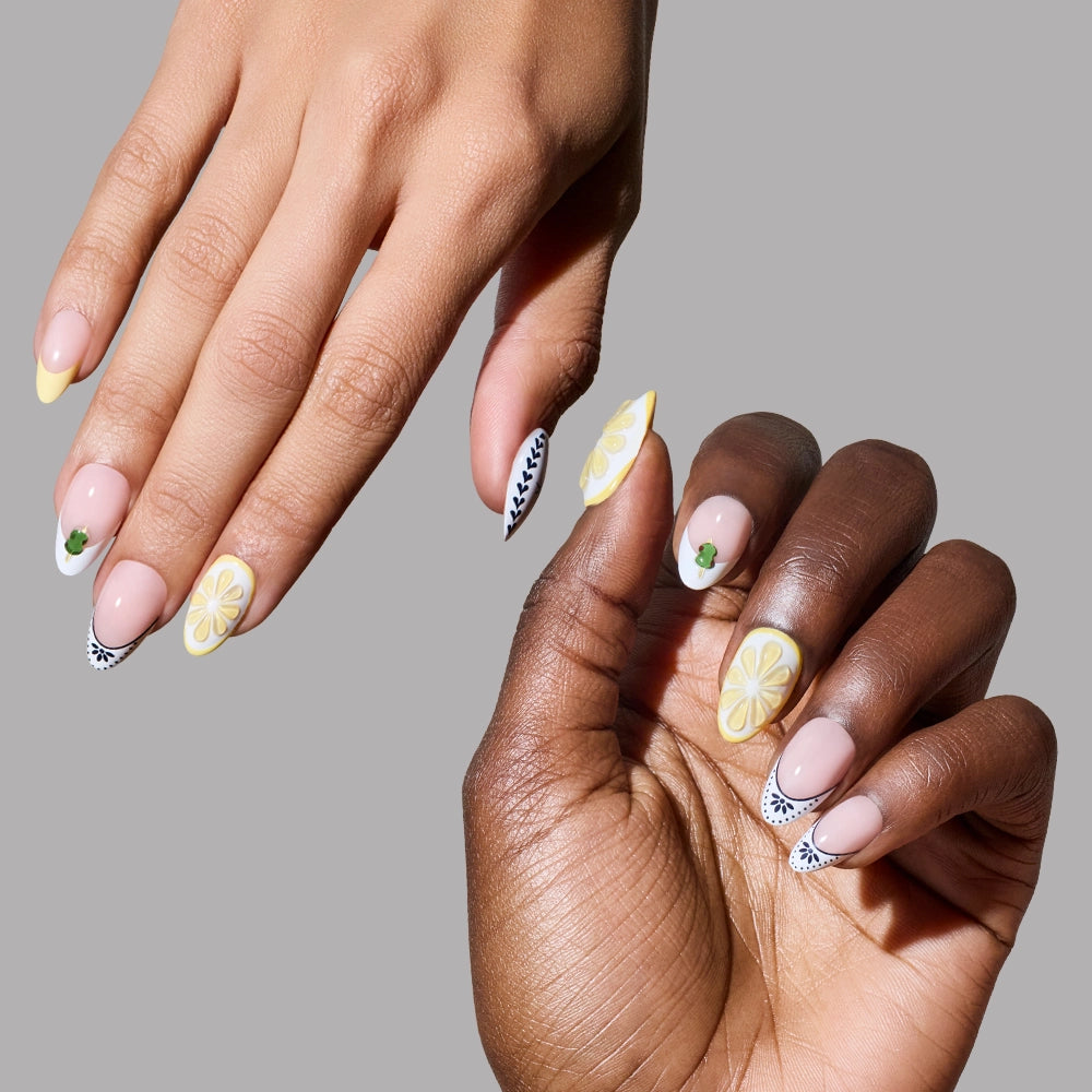 Two hands with manicured short almond nails, wearing Glamnetic’s Aperitivo Hour press-ons in yellow tips, black and white leaf designs, 3D lemon slices, and green leaves, set against a plain gray background. - Product image