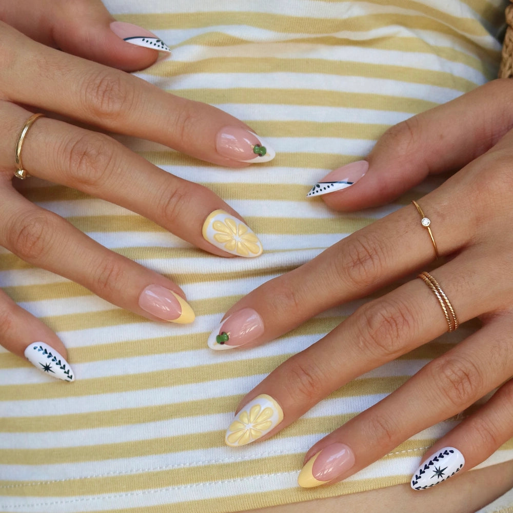 Four hands with short almond nails showcase Glamnetic's Aperitivo Hour press-ons, featuring yellow and white floral art and black dotted designs. Delicate gold rings add elegance as they rest on yellow and white striped fabric. - Product image