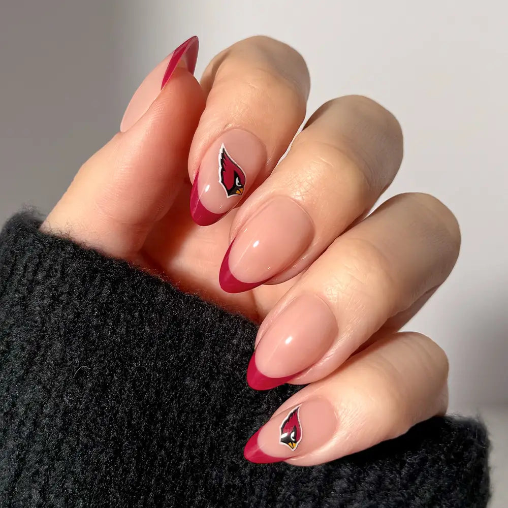 A hand showcases NFL Press-On Nails by glamnetic with almond-shaped tips in red French style and small Arizona Cardinals logos accenting two nails, set against a neutral background. The person is wearing a black, textured sleeve. - Product image