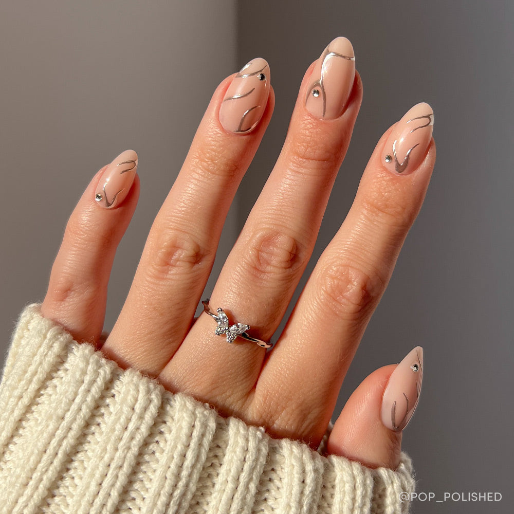A hand with short almond-shaped nude Astro nails by glamnetic, featuring abstract black lines, rhinestones, and 3D chrome swirls; adorned with a silver butterfly ring and partially covered by a cream knit sweater. - Product image