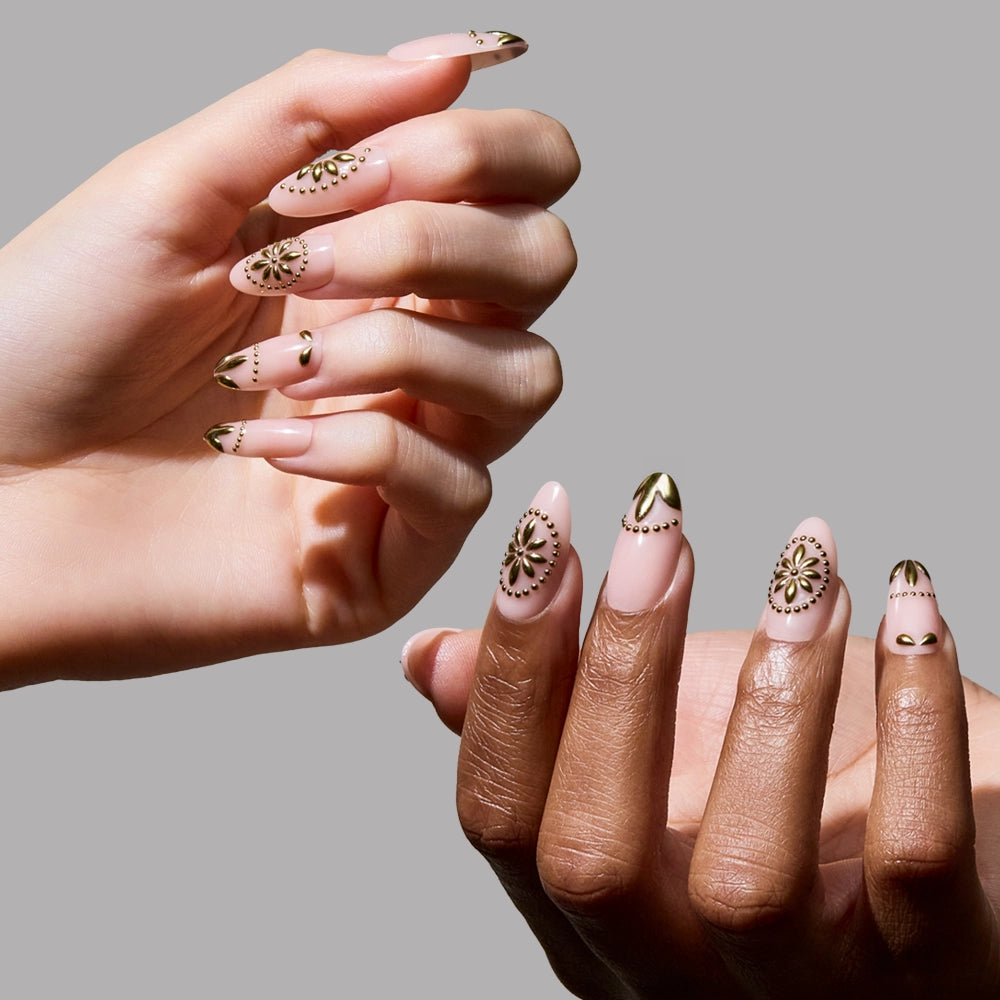 Model hands display glamnetic's Athenian Gold press-on nails, featuring long almond shapes with intricate gold geometric and floral designs on a nude base, set against a plain gray background. - Product image
