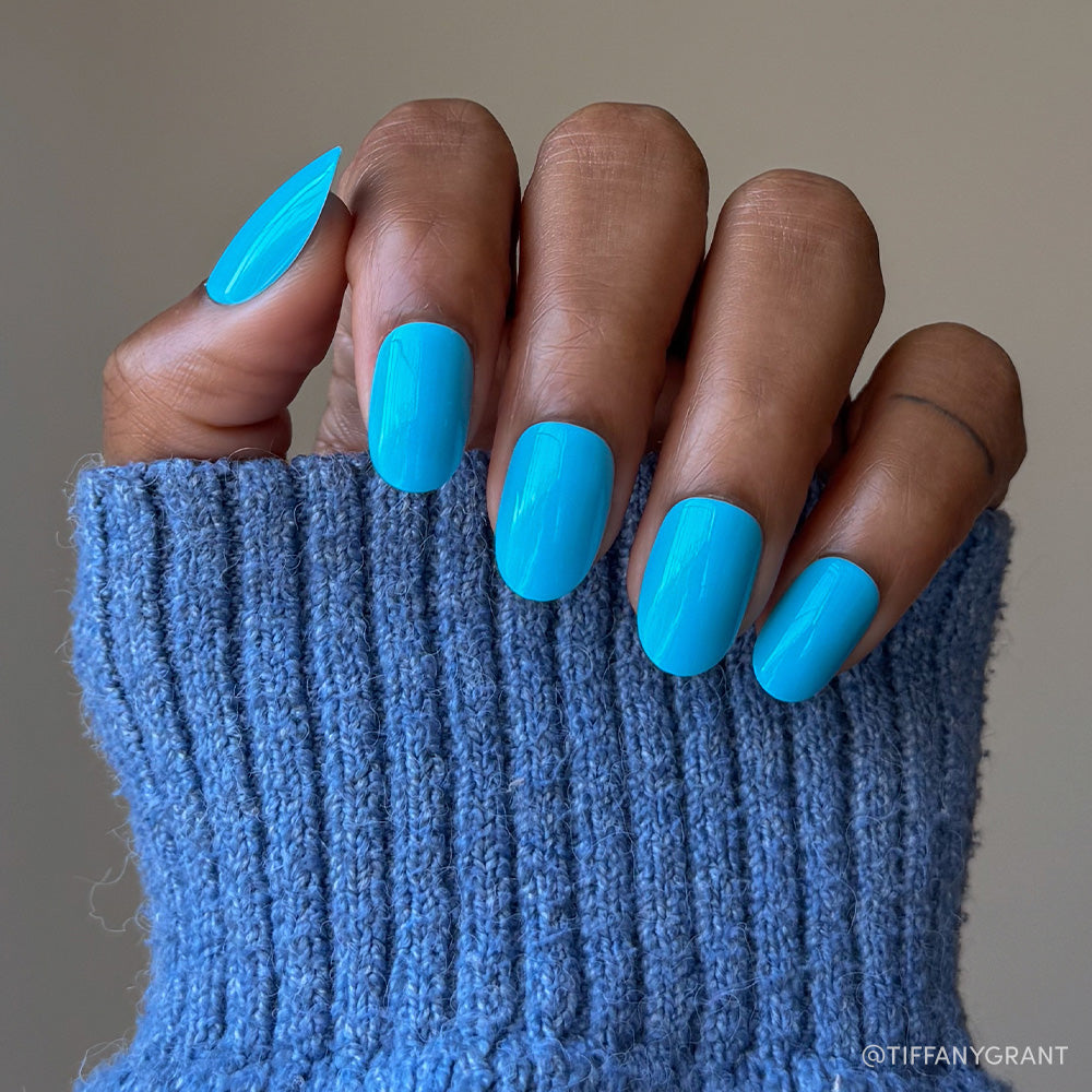 A hand with medium brown skin in a blue knit sweater displays neatly manicured nails painted with glamnetic’s Azure Blue, perfect for summer fun and long-lasting wear. - Product image