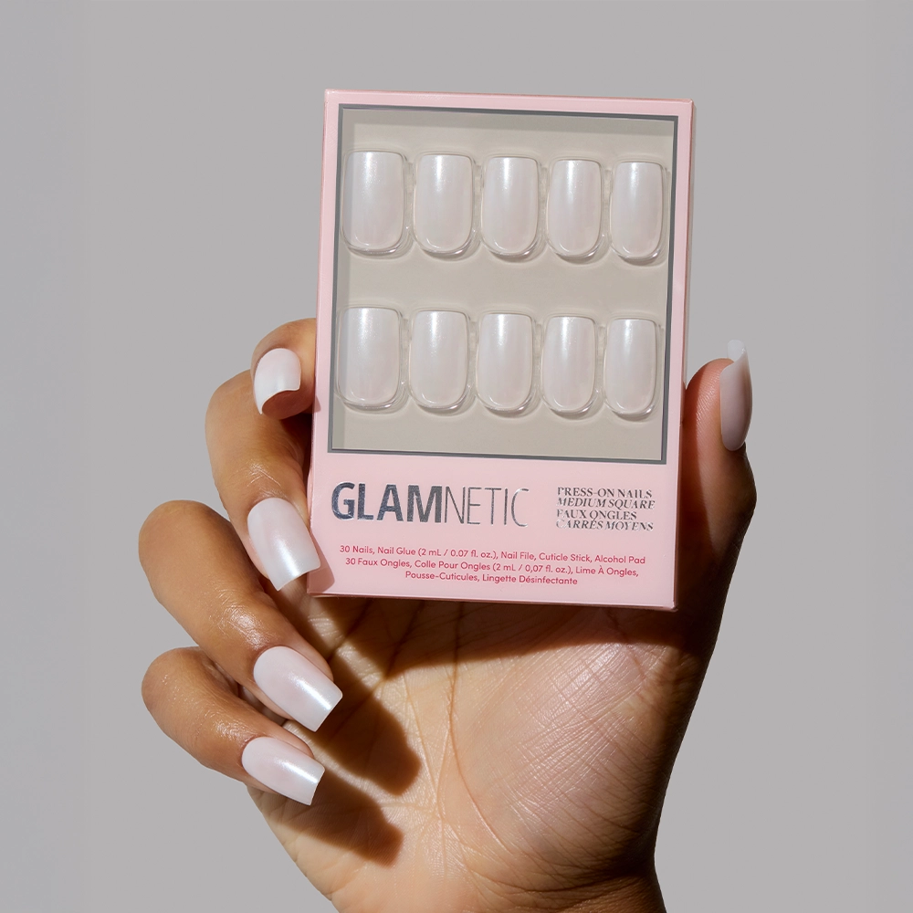 A hand with glossy, neatly manicured medium square nails holds a box of glamnetic Bailey vegan press-on nails against a neutral background. The package shows ten press-on nails arranged in two rows. - Product image