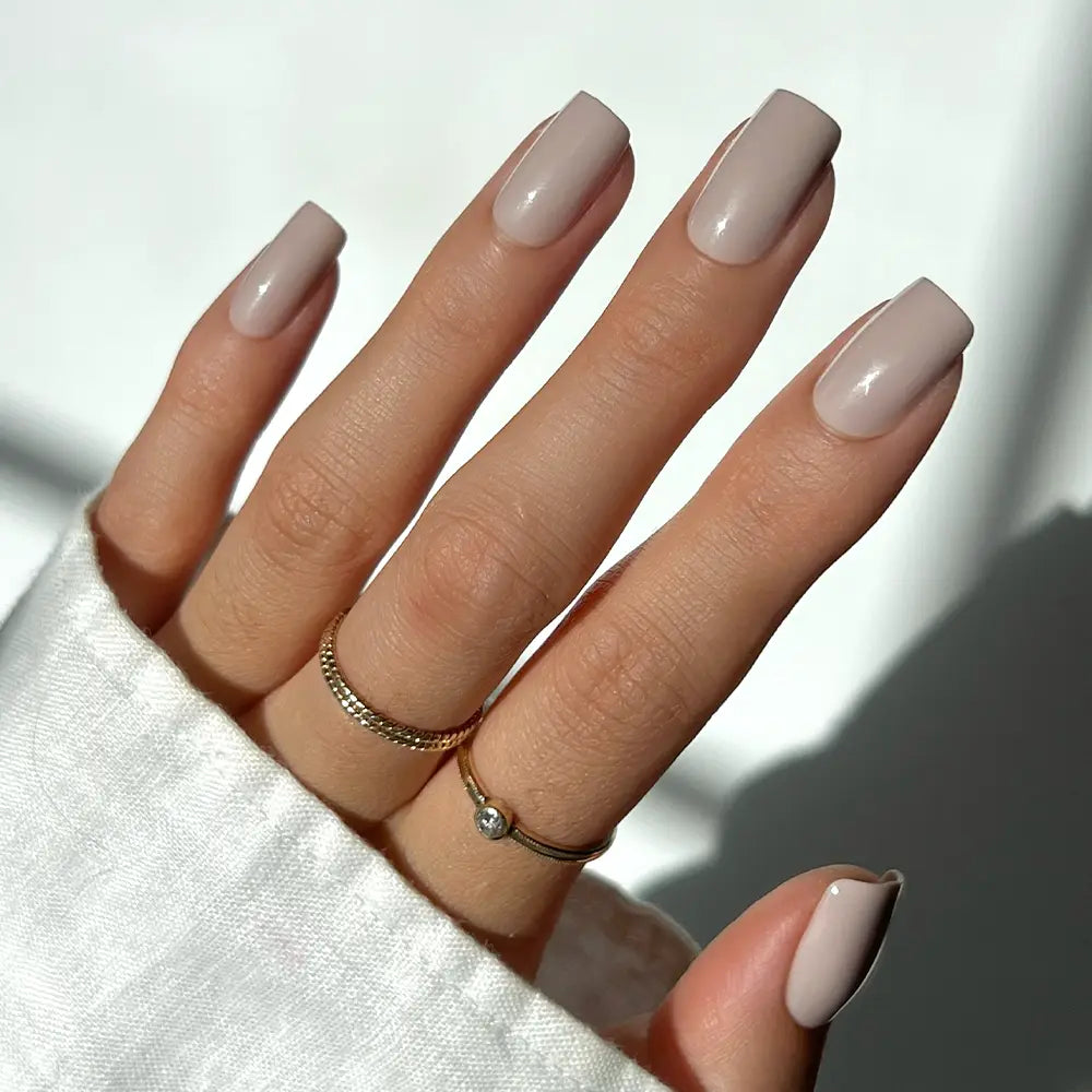 A hand with neatly manicured, glossy nude glamnetic Bare Bliss press-on nails in a medium square shape, wearing two gold rings, is shown against a bright background. The person is dressed in a white long-sleeve shirt. - Product image