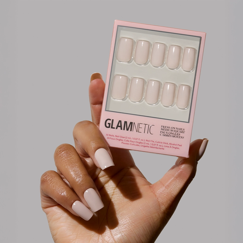 A hand with neatly manicured, light pink nails holds a box of glamnetic Bare Bliss press-on nails in a matching nude shade, highlighting polished simplicity against a plain gray background. - Product image