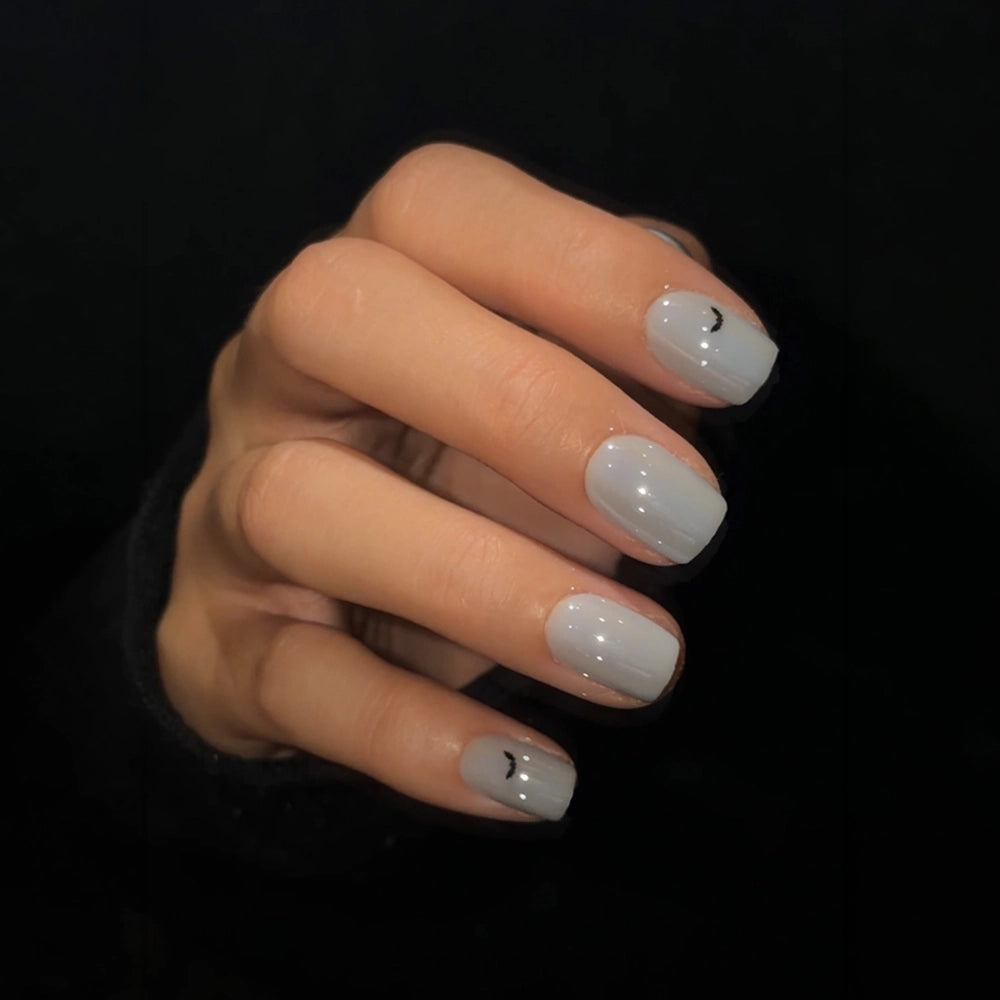 A close-up of a hand with short, squoval press-on nails painted in a glossy, light grey polish against a black background. Each nail features a tiny, minimalist black crescent moon design near the cuticle. - Product image