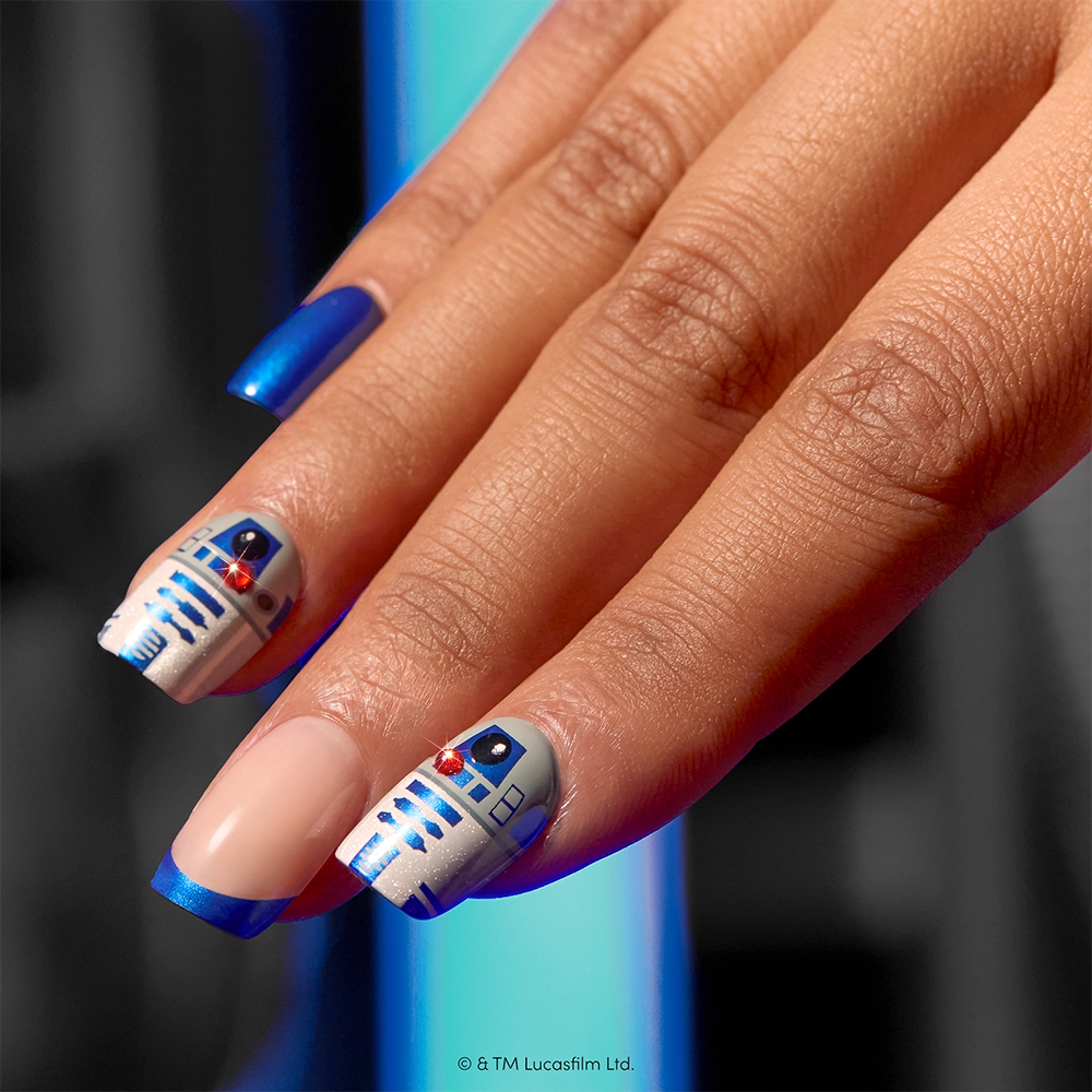 A hand featuring the Beep-Boop press-on nails by glamnetic, adorned with blue and nude nail art inspired by R2-D2 from Star Wars. These short squoval STAR WARS press-ons display intricate droid patterns on select fingers, while others are painted in solid blue or nude with bold blue tips. Perfect for fans, Beep-Boop combines playful detail and galactic flair in an easy-to-wear shape. - Product image