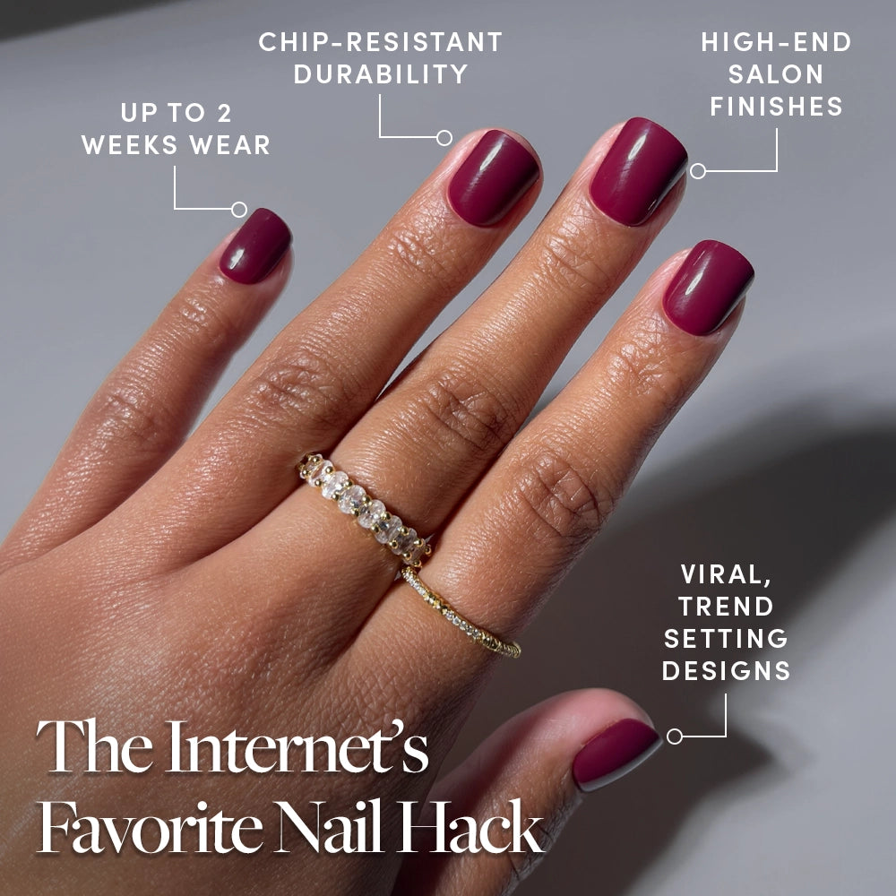 A hand with neatly manicured, super short maroon "Berry Baby" nails by glamnetic, featuring up to 2 weeks wear, chip-resistant durability, salon-quality finish, and viral trend-setting press-on designs. Two gold rings accent the look. - Product image