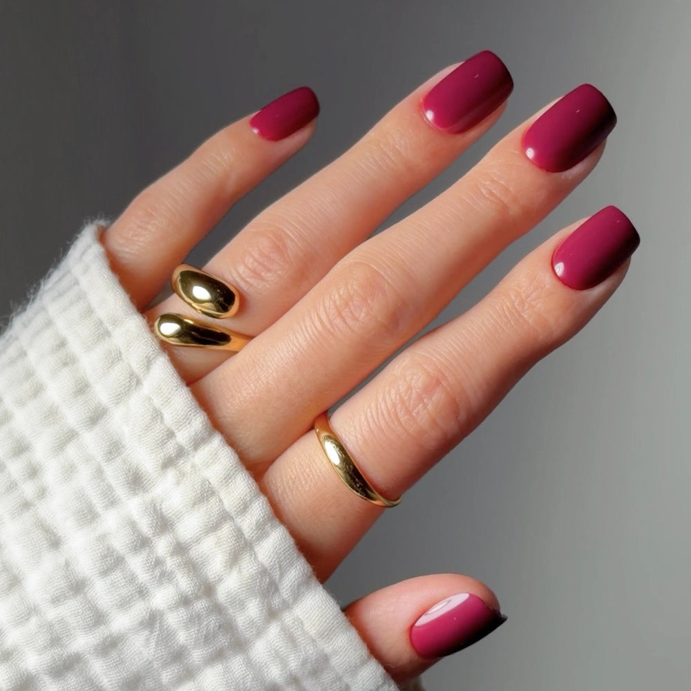 A hand with neatly manicured, glossy, short squoval nails in glamnetic's Berry Crush—a deep pink shade—wears two shiny gold rings. The person is dressed in a white textured long-sleeve top with a softly blurred background.