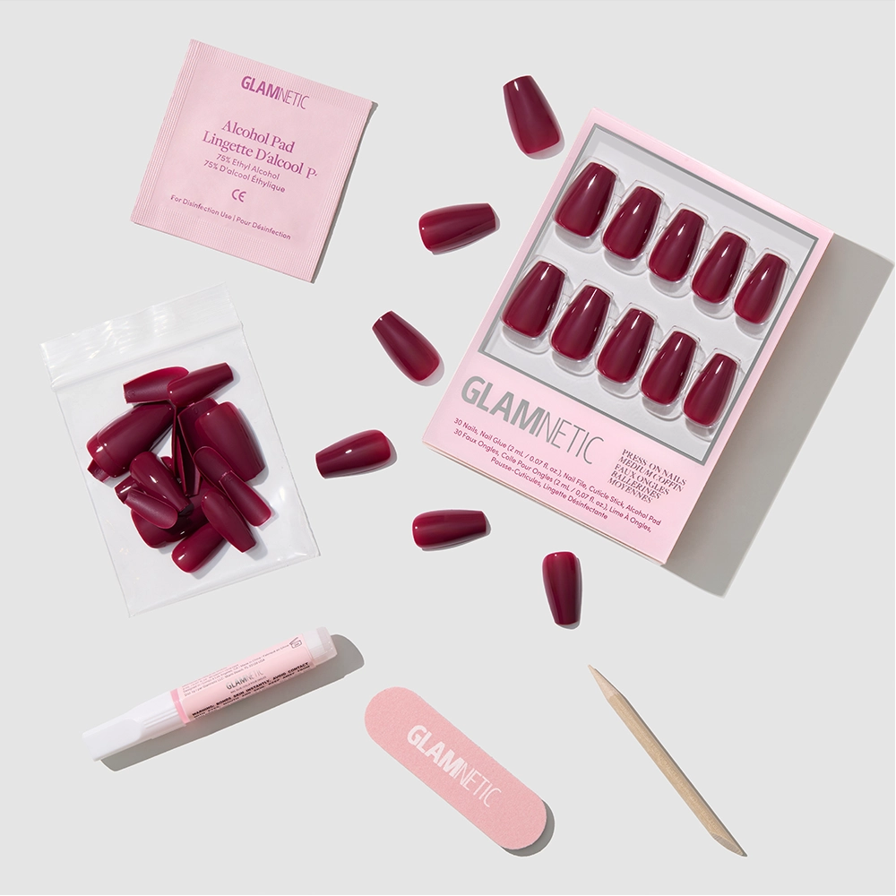 The Berry Fierce set by glamnetic includes maroon coffin-shaped press-on nails, extra nails, an alcohol pad, nail glue, nail file, and wooden cuticle stick—neatly arranged with pink packaging labeled “glamnetic.”.