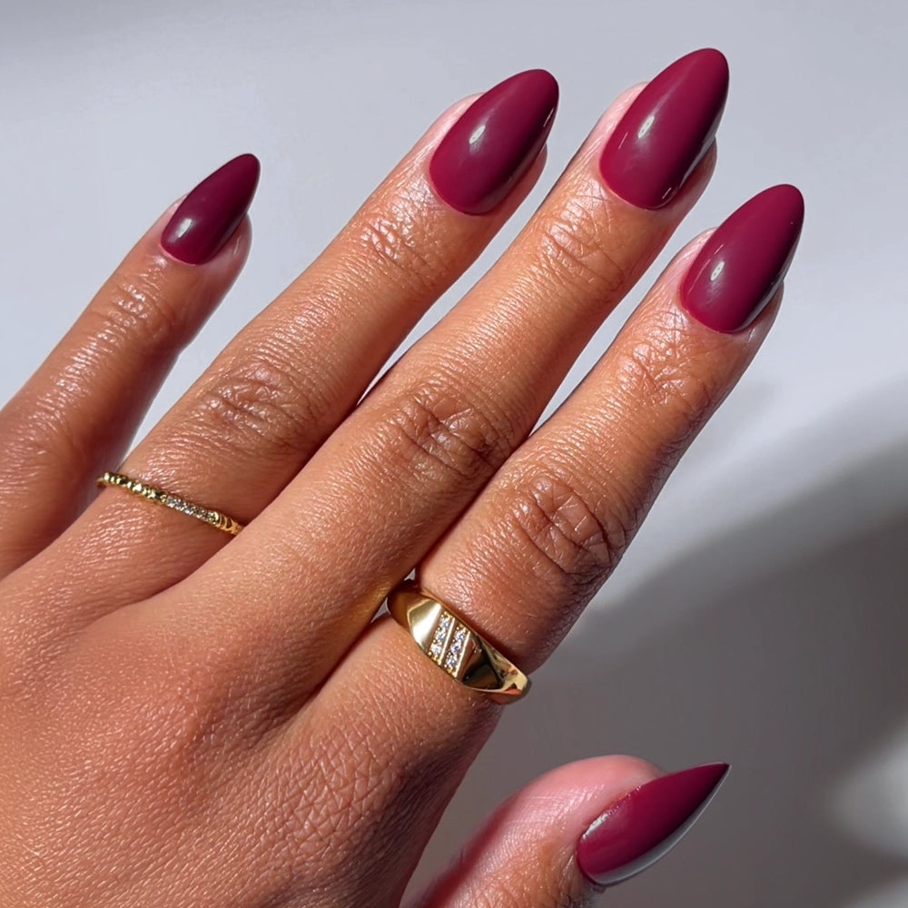 A hand with short almond nails features glamnetic’s Berry Kiss, glossy burgundy press-ons. A gold geometric ring and thin twisted band add chic flair, while a softly blurred background enhances the elegant vibe.