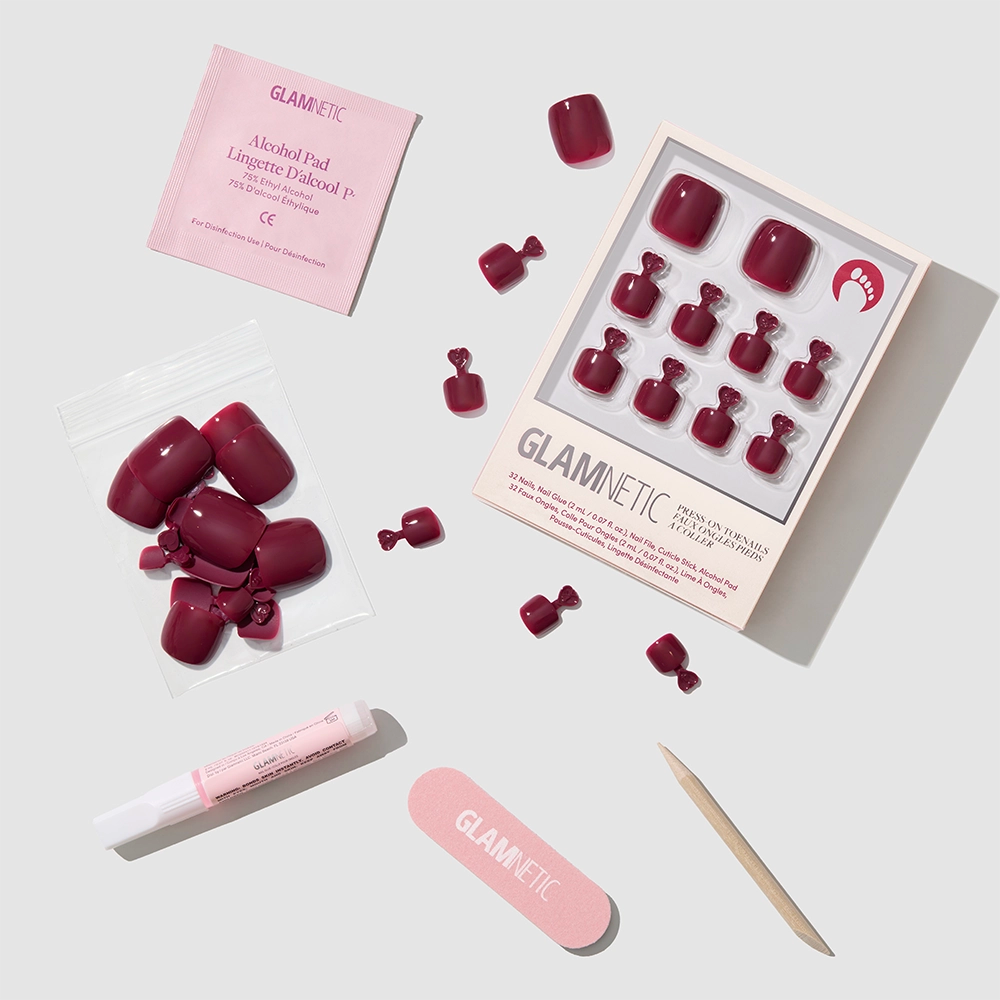 The glamnetic Berry Maroon Toenail set, featuring glossy press-on toenails, a pink nail file, wooden cuticle stick, nail glue, alcohol pad, and extra nails, is arranged on a light grey background for effortless style in minutes. - Product image