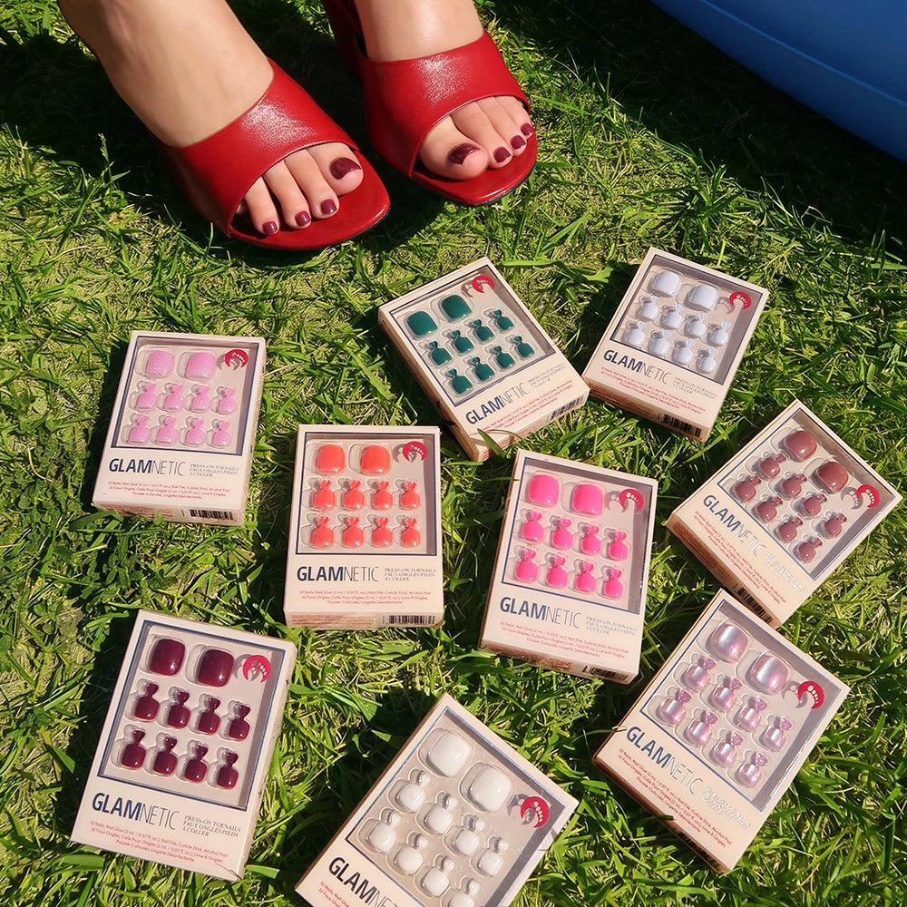 A person with painted toenails in red sandals stands on grass, surrounded by ten glamnetic press-on toenail boxes in assorted styles and shades, including the vegan, cruelty-free Berry Maroon Toenail option. - Product image