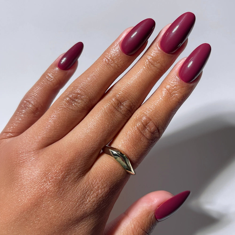 A hand with medium skin tone displays long, medium oval nails painted in glamnetic’s Berry Tempted matte wine-red shade, wearing a gold ring with a marquise green stone on the middle finger, set against a plain white background.