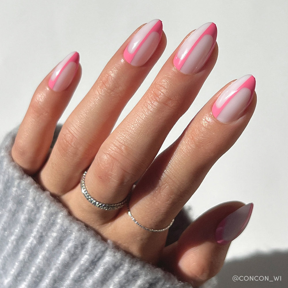 A hand models glamnetics Better Half nails—short almond shape with a milky white base and pink French tips with translucent centers, accented by modern abstract lines. Two thin silver rings and a light gray ribbed sweater complete the look against a simple background.