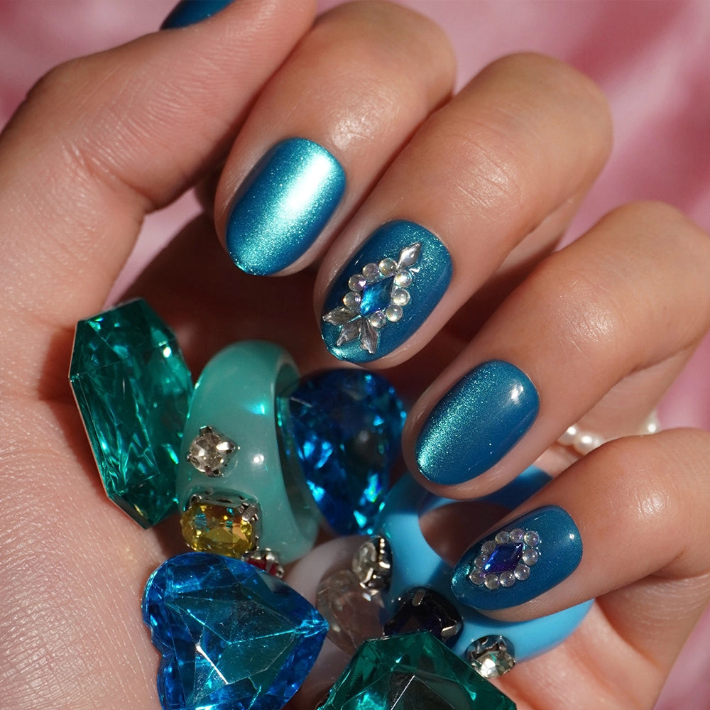 A hand with shimmery teal blue nails—one accentuated with rhinestones and gems—holds blue and green gemstone-like decorations against a soft pink background, beautifully highlighting the Blue Topaz short oval press-on nails by glamnetic for an effortlessly chic look. - Product image