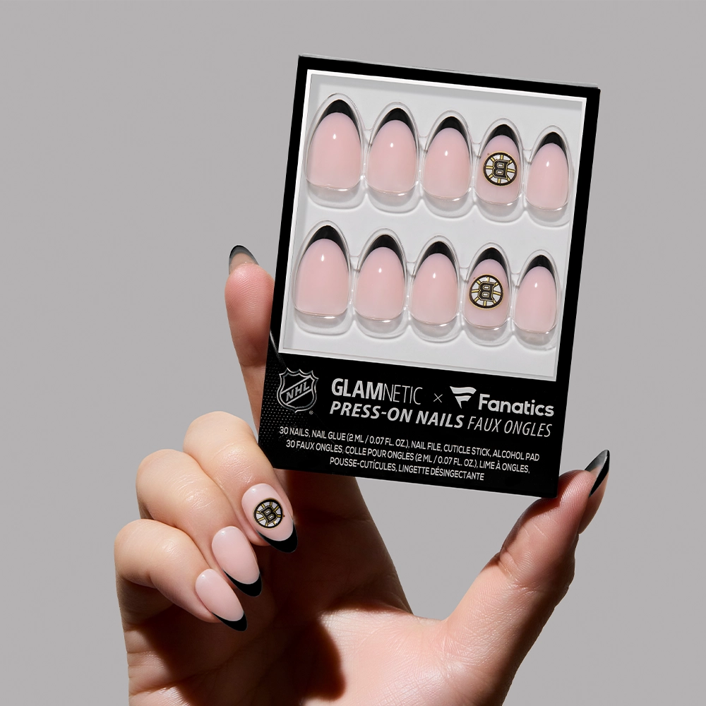 A hand with black-tipped press-on nails holds a box of glamnetic Boston Bruins Press-On Nail Set. The light pink nails feature black French tips and a gold sports-themed design, perfect for showcasing team spirit.