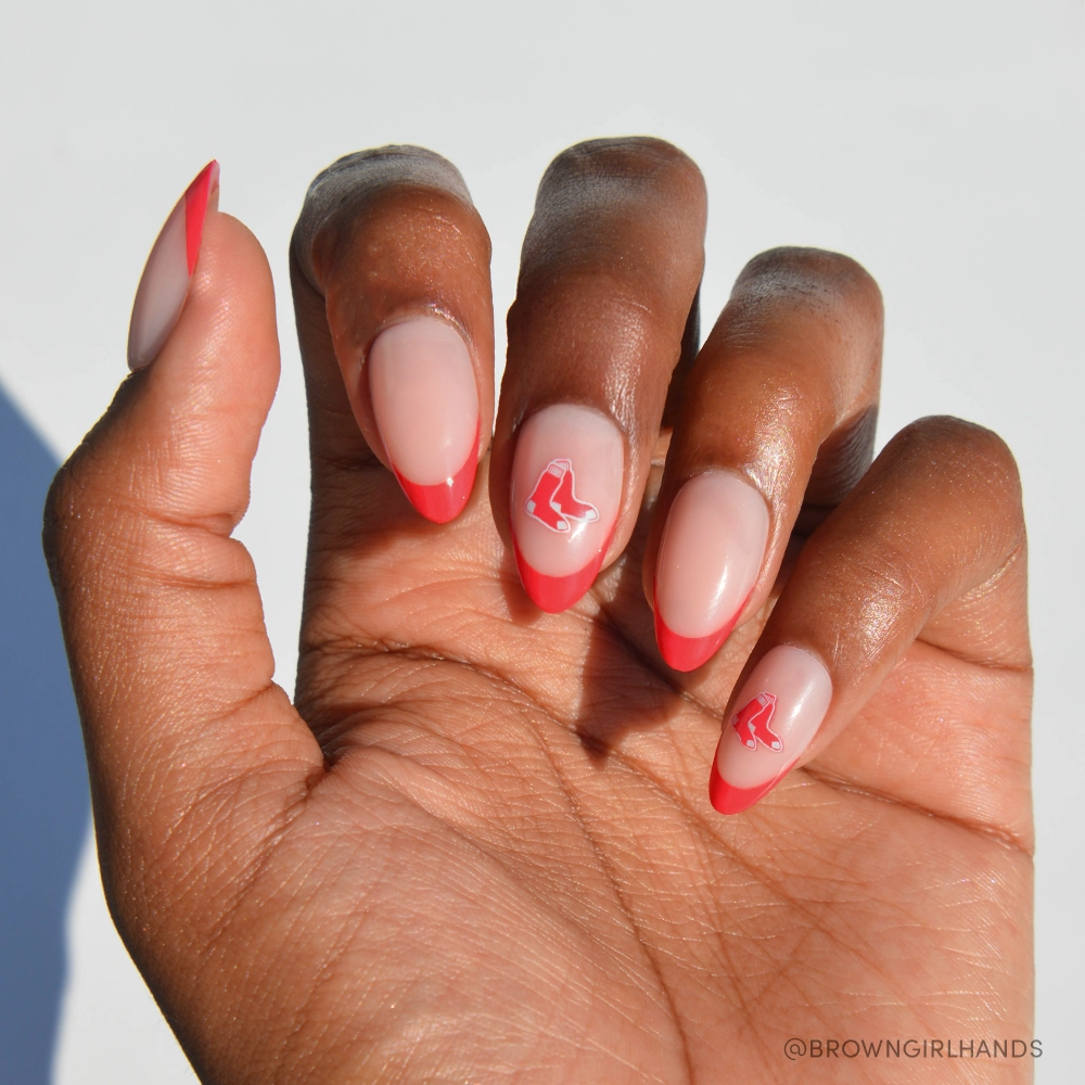 A hand with almond-shaped nails features glamnetics Boston Red Sox Press-On Nail Set, displaying red French tips and a small red sock graphic on the middle and ring fingers against a white background. - Product image