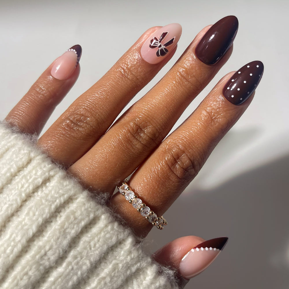 Glamnetic Bow Season short almond nails featuring brown and nude designs, white dots, a black bow accent, and chocolate brown French tips with a white dot border.