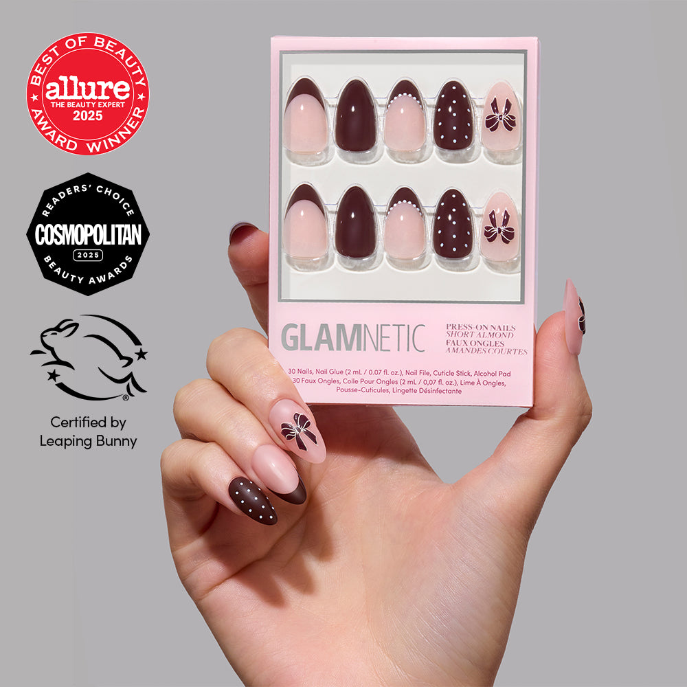 A hand holds a Bow Season box of chocolate brown and light pink short almond-shaped press-on nails with bow and polka dot designs, displayed next to three award seals from Allure and Cosmopolitan.