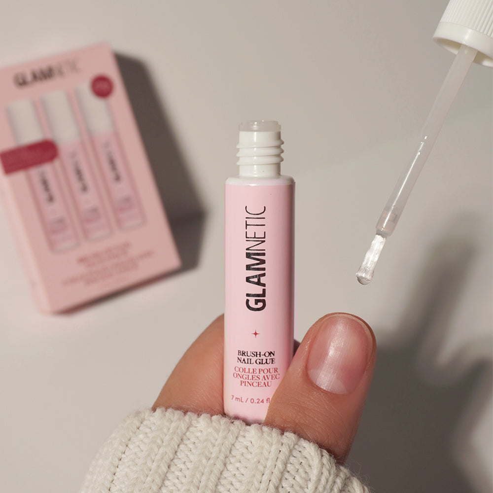 A hand holds a pink tube from the glamnetic Glue Trio with the cap off, revealing a brush applicator. Ideal for applying press-on nails and achieving salon-quality results at home, this nail glue comes in stylish packaging; in the background, a blurred pink box with product graphics can be seen. - Product image