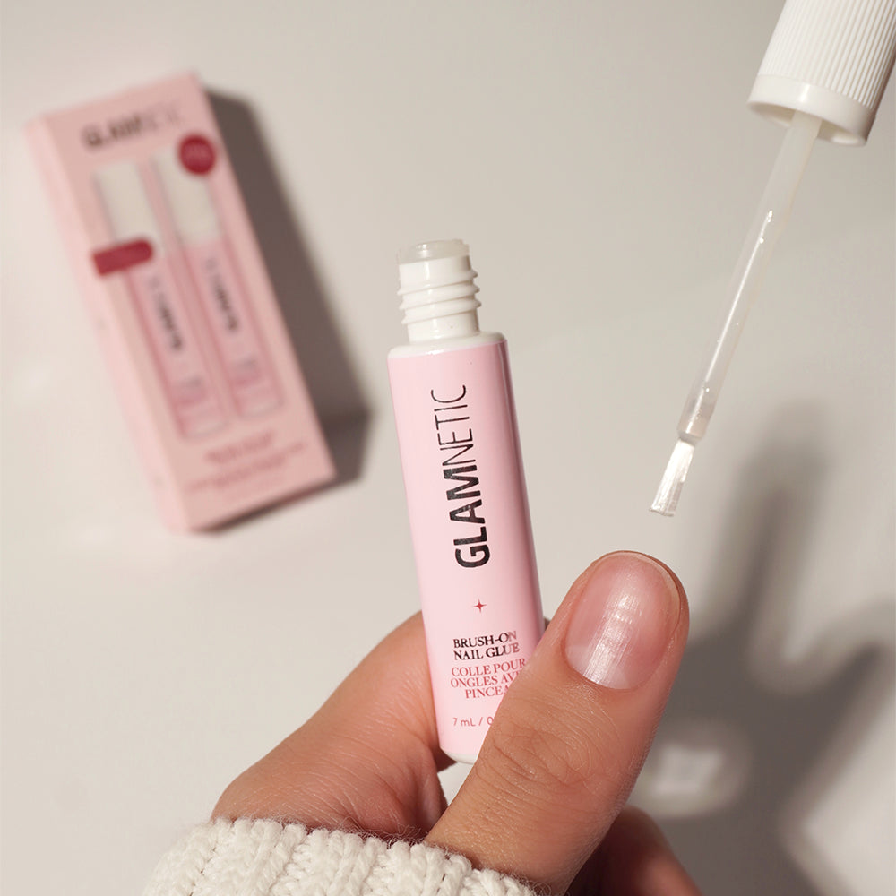 A hand holds a pink glamnetic Glue Duo tube with the brush applicator removed—an ideal prep step for applying your press-on nails. In the background, a matching pink product box rests on a light surface, suggesting salon-quality results from the comfort of home. - Product image