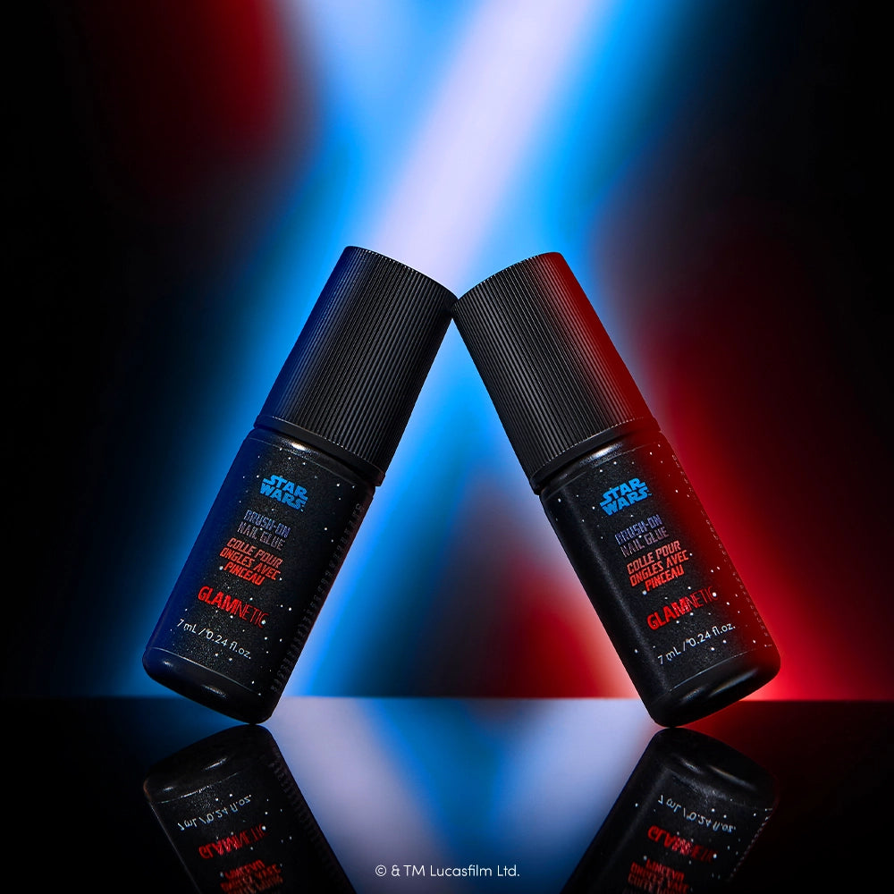 STAR WARS™ | Glamnetic Brush-On Nail Glue - Two STAR WARS™ | Glamnetic Brush-On Nail Glue bottles stand upright, angled toward each other against a dramatic backdrop of blue and red light beams. Each bottle features a sleek black cap with eye-catching red and blue text, perfectly matching the glamnetic STAR WARS™ collection for fans who want to coordinate their fast-drying nail glue with their personal care essentials.